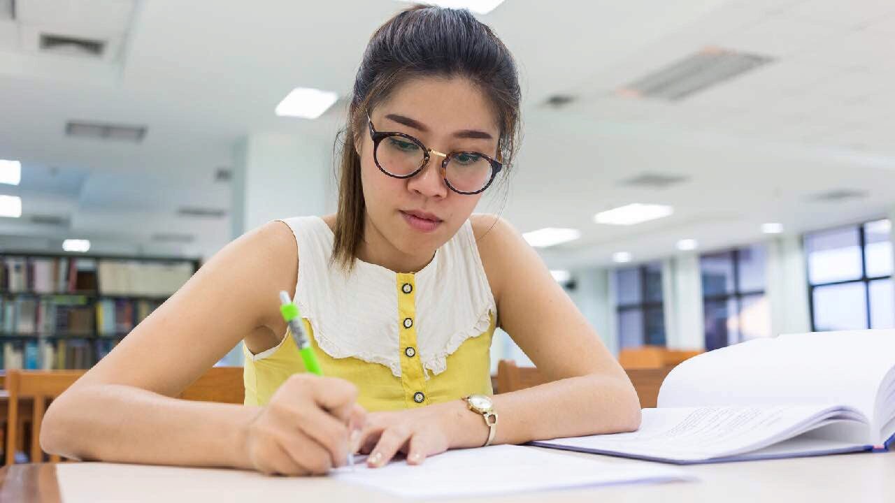 A person wearing glasses is studying and writing in a book while seated at a table in a library.