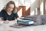 A person writes in a notebook at a table, with a gray backpack and open binder nearby. A glass of water is on the table.