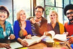 A diverse group of five people smiling and sitting at a table with open books, suggesting a collaborative study session or group discussion.