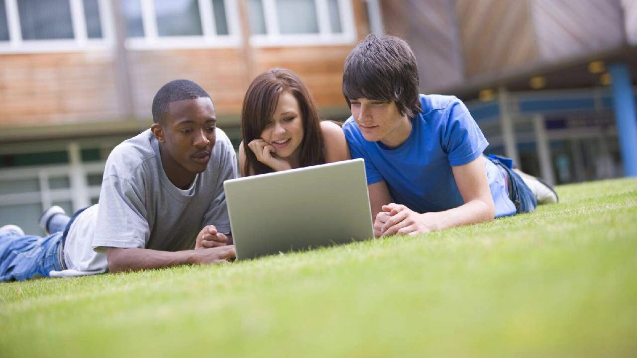 Three people are lying on grass, looking at a laptop. They appear engaged and relaxed, suggesting a setting of outdoor study or leisure.