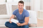 A person sitting on a living room floor, smiling and holding a piece of paper, with a white sofa and cushions in the background.
