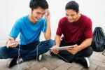 Two people sitting on the ground, one wearing headphones connected to a smartphone, the other holding a tablet. Both are smiling and appear engaged.