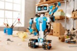 A colorful toy robot built with interlocking bricks on a wooden table, surrounded by crafting materials and containers of loose bricks, in a well-lit room.