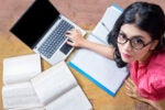 A person wearing glasses and a pink shirt is sitting on the floor, surrounded by books and a laptop, appearing to study or work.