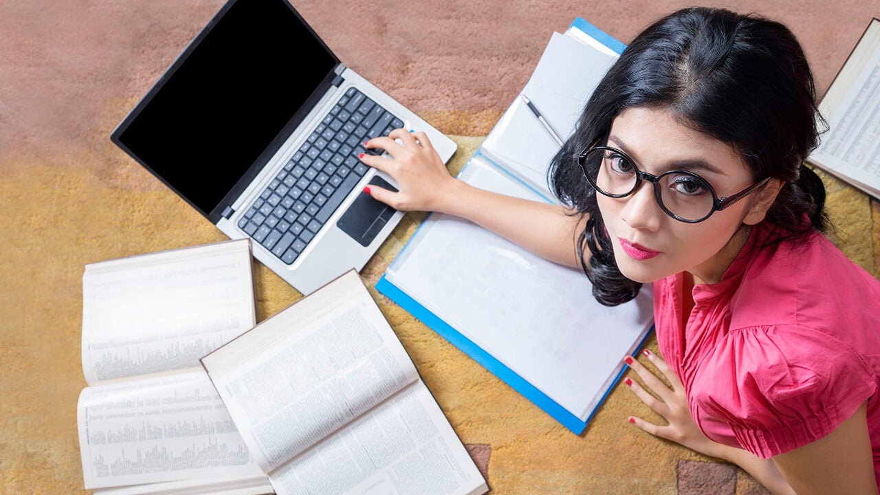 A person wearing glasses and a pink shirt is sitting on the floor, surrounded by books and a laptop, appearing to study or work.