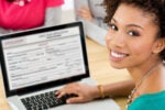 A person smiles at the camera while using a laptop, which displays an application form.