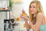 A person with long blond hair enjoys a bowl of cereal with milk in a kitchen setting, next to a coffee maker and various kitchen items.