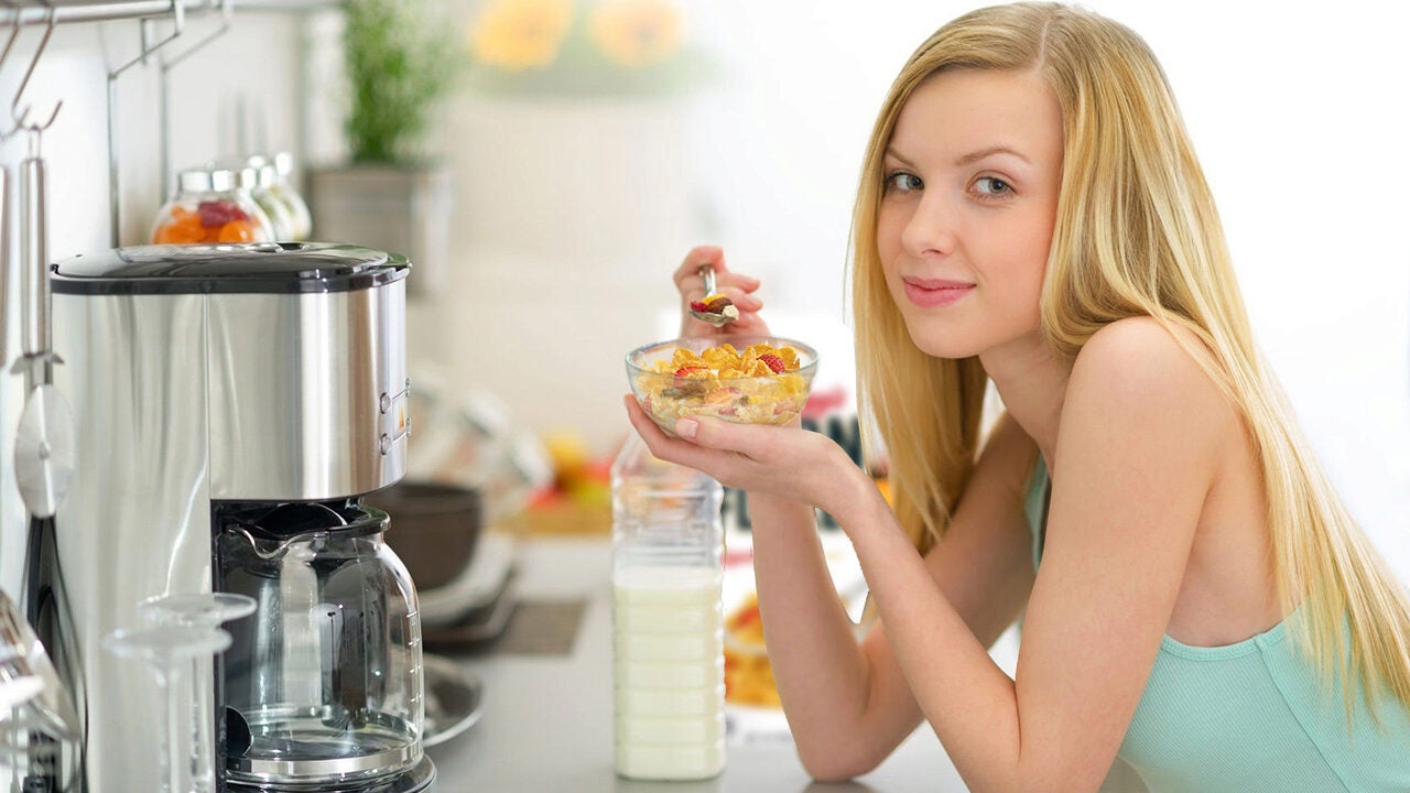 A person with long blond hair enjoys a bowl of cereal with milk in a kitchen setting, next to a coffee maker and various kitchen items.