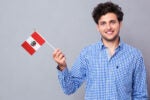 A person smiling while holding a small Peruvian flag, wearing a blue checkered shirt against a neutral gray background.