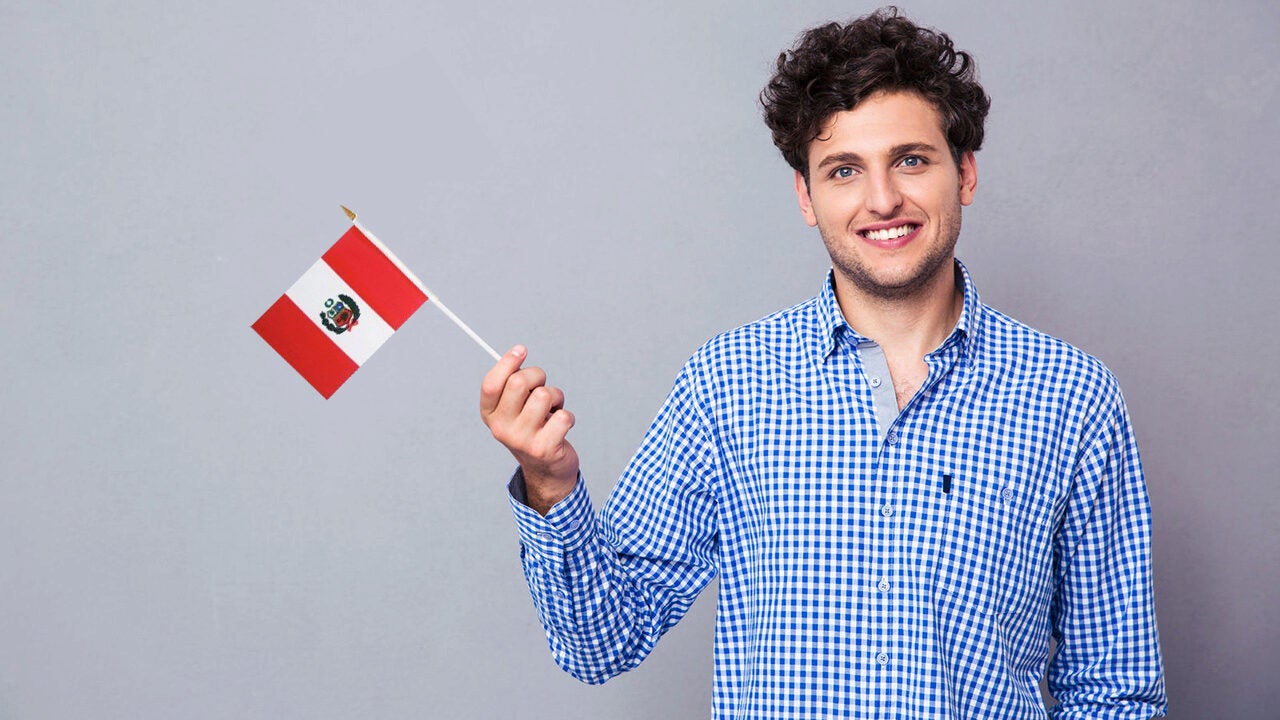 A person smiling while holding a small Peruvian flag, wearing a blue checkered shirt against a neutral gray background.