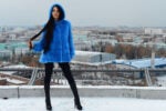 A person wearing a bright blue fur coat and hood stands on a snowy rooftop with a cityscape in the background.