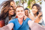 A group of five young people taking a selfie together outdoors, smiling and having fun. Palm trees in the background suggest a sunny, tropical location.