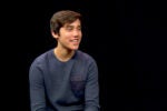 A person smiling while sitting against a plain black background, wearing a dark blue long-sleeve shirt.