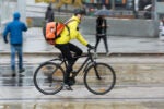 A cyclist wearing a yellow jacket and helmet rides a bike with an orange backpack in a rainy urban setting. Pedestrians with umbrellas are in the background.