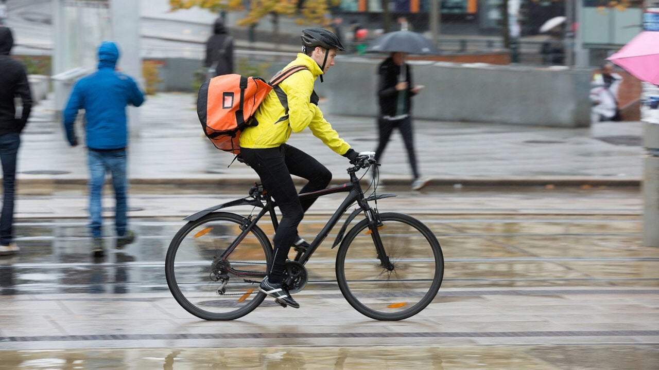 A cyclist wearing a yellow jacket and helmet rides a bike with an orange backpack in a rainy urban setting. Pedestrians with umbrellas are in the background.