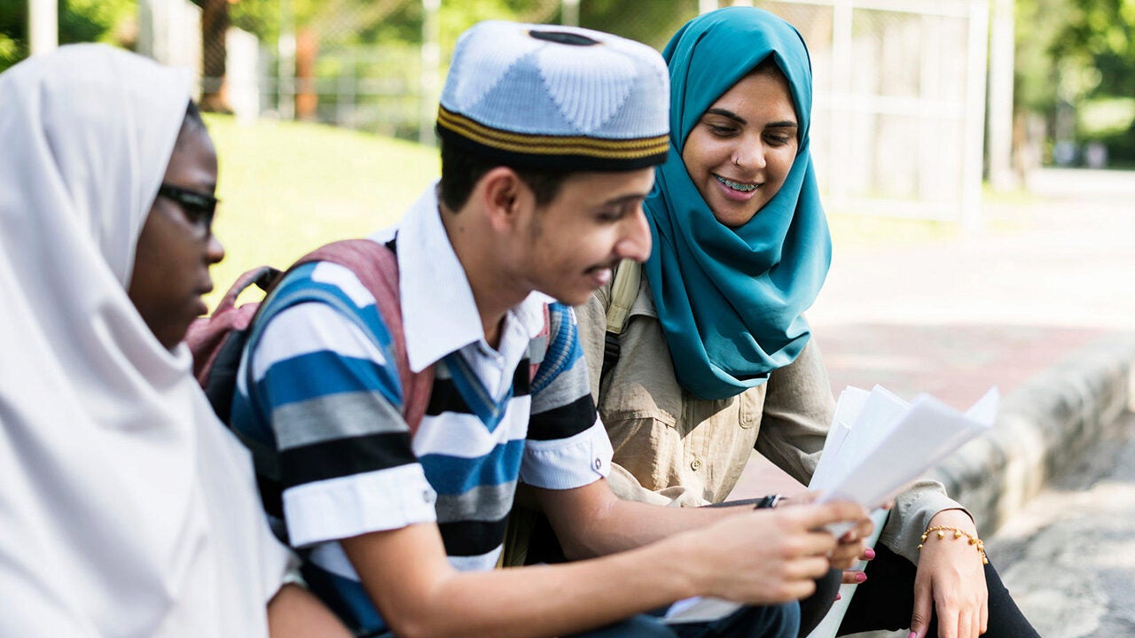 Three people sitting together, two wearing headscarves and one wearing a hat, looking at documents in a casual outdoor setting.