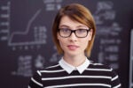 Headshot of a person wearing glasses and a black and white striped shirt, standing in front of a blackboard with charts and graphs.