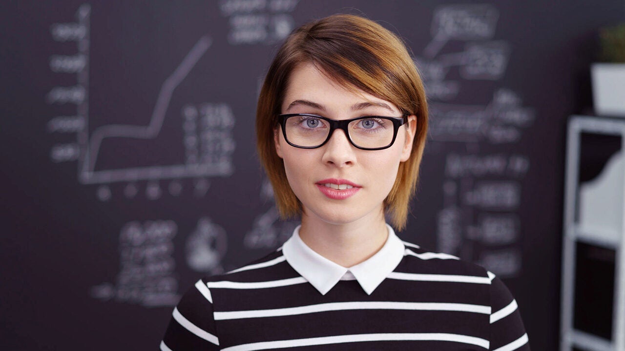 Headshot of a person wearing glasses and a black and white striped shirt, standing in front of a blackboard with charts and graphs.