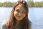 A person smiling outdoors near a body of water, with trees in the background.