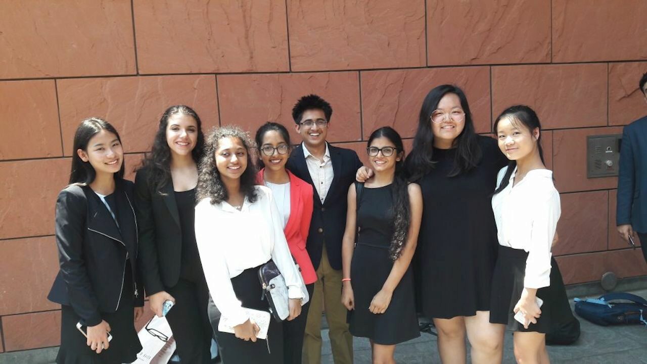 A group of eight people standing together in business attire, smiling, with a reddish-brown tiled wall in the background.