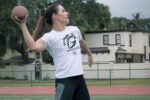 A person on a sports field is throwing a football, wearing a white shirt with a flag football camp logo. Trees and a building are in the background.