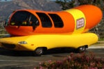 A car shaped like a giant hot dog, known as the Wienermobile, with "Oscar Mayer" branding on the side, parked in a scenic area.