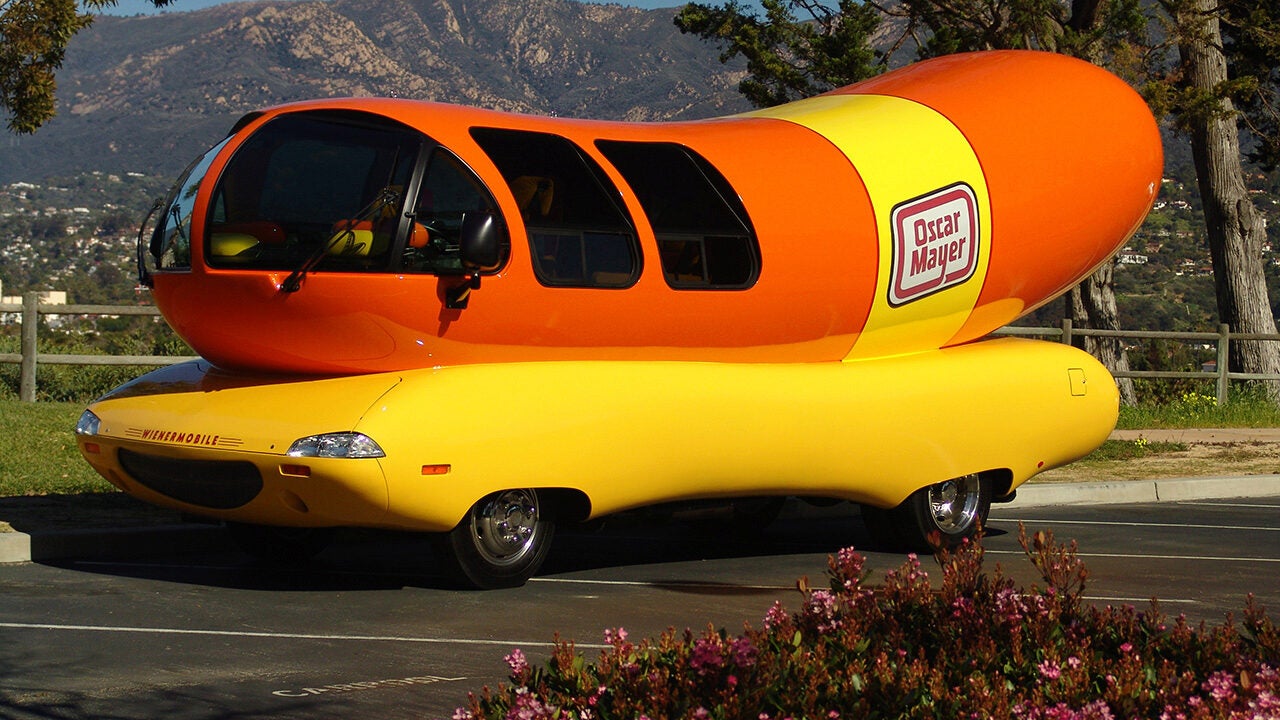 A car shaped like a giant hot dog, known as the Wienermobile, with "Oscar Mayer" branding on the side, parked in a scenic area.