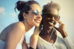 Two smiling people wearing sunglasses, enjoying music with shared earphones outdoors on a sunny day.