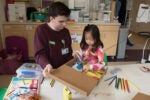 An adult and a child engage in a crafting activity at a table with colored pencils, paper, and art supplies in a classroom or hospital setting.