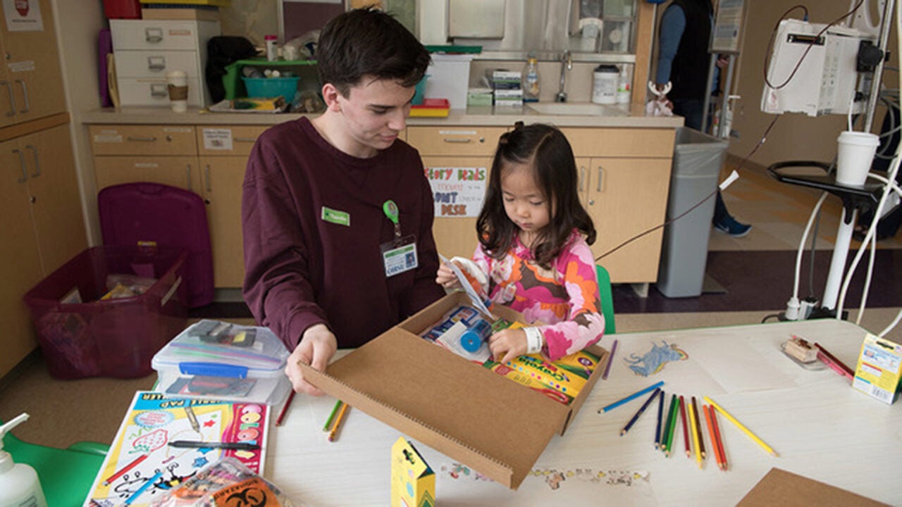 An adult and a child engage in a crafting activity at a table with colored pencils, paper, and art supplies in a classroom or hospital setting.