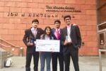 Four young people in business attire hold a large check and two trophies, standing in front of a brick building labeled "The Wharton School, University of Pennsylvania, Jon M. Huntsman Hall."