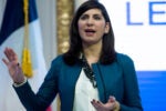 A person is speaking and gesturing, wearing a teal jacket. Behind them is a flag and a partially visible sign with the word "LEAD."