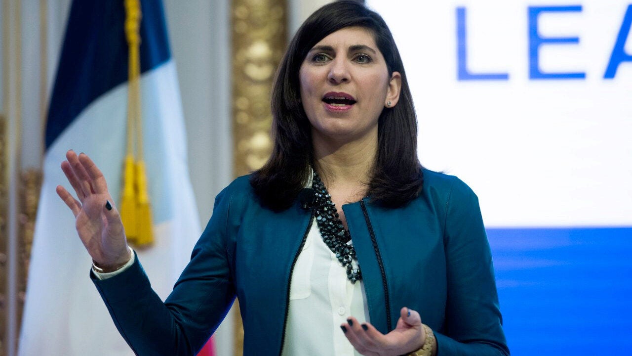 A person is speaking and gesturing, wearing a teal jacket. Behind them is a flag and a partially visible sign with the word "LEAD."