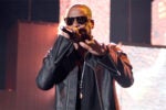 A person wearing sunglasses and a leather jacket is performing on stage, holding a microphone.