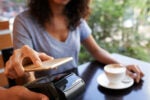 A person using a smartphone to make a contactless payment on a terminal in a café setting, with a cup of coffee on the table.