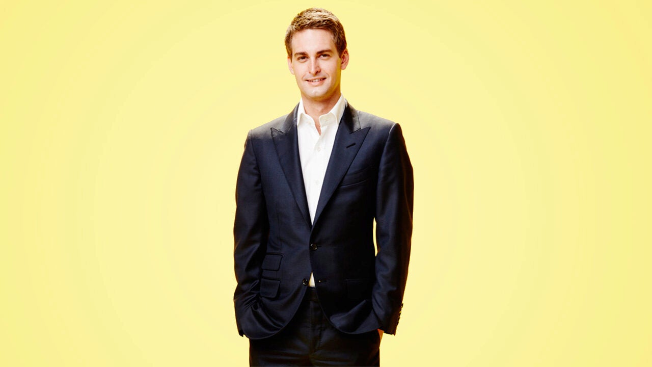 A person in a dark suit and white shirt standing against a yellow background, smiling at the camera.