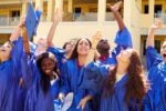 A group of graduates wearing blue caps and gowns celebrate by raising their arms and tossing their caps into the air.