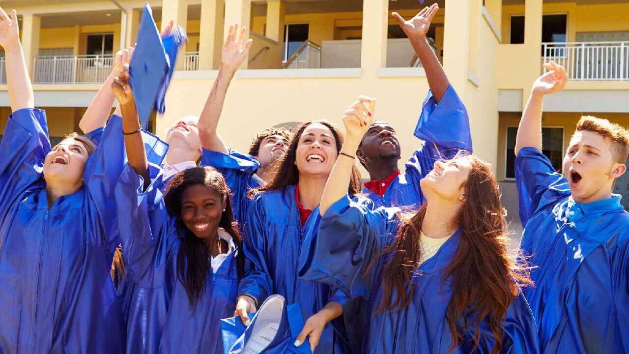 A group of graduates wearing blue caps and gowns celebrate by raising their arms and tossing their caps into the air.