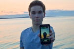 A person standing by a body of water at sunset, holding a smartphone displaying an app called SmartLens that identifies objects.