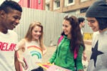 A group of four young people standing outdoors, smiling and looking at colorful papers in their hands. The setting appears urban, with buildings and construction fencing in the background.