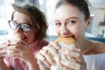 Two people joyfully eating burgers, one wearing glasses and the other in a light-colored top, sitting inside a bright, casual setting.