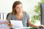A person smiling while working at a desk, holding documents with colorful charts. There is a stack of papers beside them and a computer monitor in view.