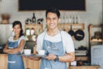 Two people in aprons smiling in a cozy café setting, with one holding a tray with coffee cups.