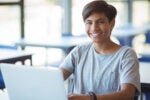 A person sitting at a desk smiling and working on a laptop in a bright room.