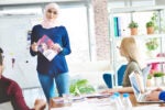 A person in a hijab stands in front of a group in a bright office, holding a magazine and presenting while others listen or take notes.