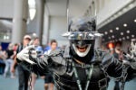 Person in a black leather costume with spiked headgear and chains, posing with open mouth and outstretched hands, at a convention.