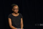 A headshot of a person wearing glasses and a black T-shirt, sitting against a dark background with the letters KWHS in the corner.