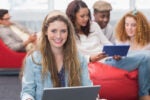 A group of young adults are interacting in a casual space. In the foreground, a person is smiling while using a laptop. Others in the background use a tablet and chat.