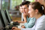 A group of young adults interacting and working at a row of computer monitors, suggesting a collaborative learning or team project environment.