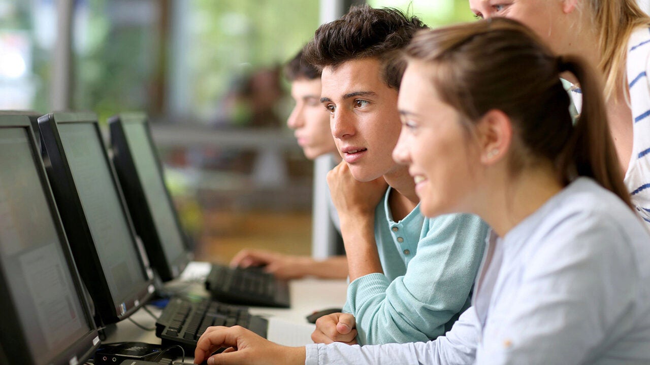 A group of young adults interacting and working at a row of computer monitors, suggesting a collaborative learning or team project environment.